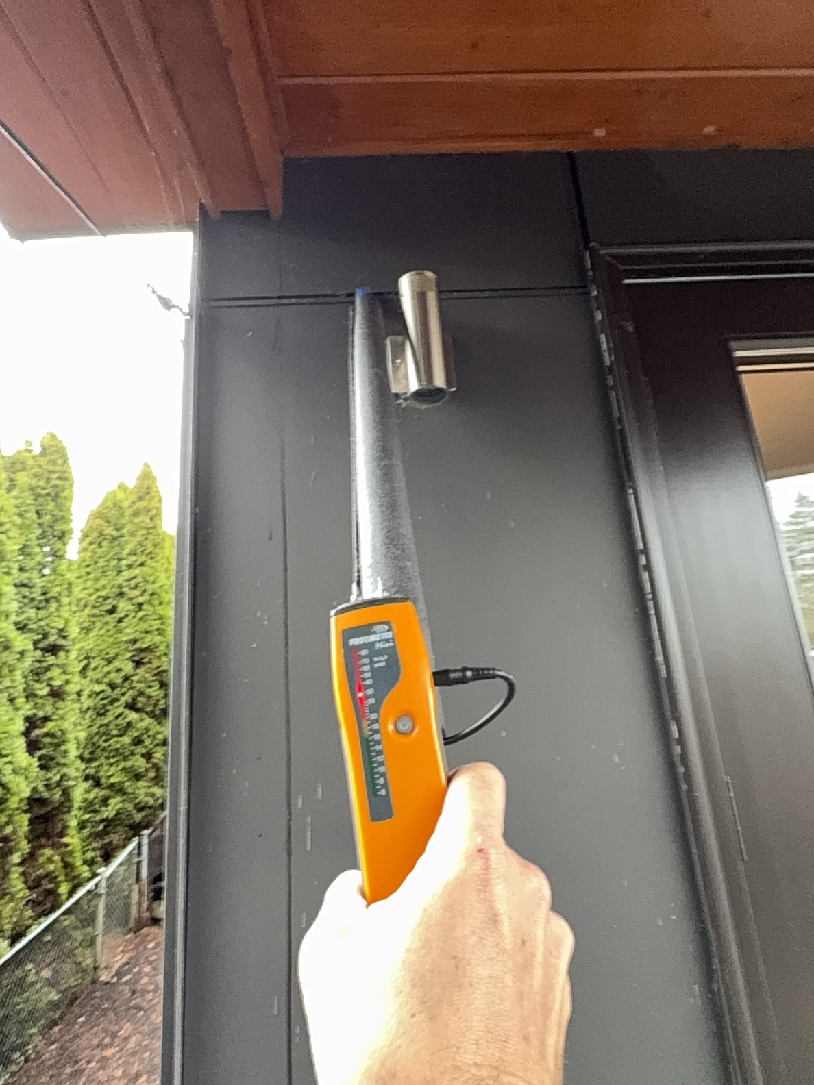A moisture meter held to the wall directly beneath the deck shows the needle pegged in the red zone — confirming water has penetrated through the surface above.